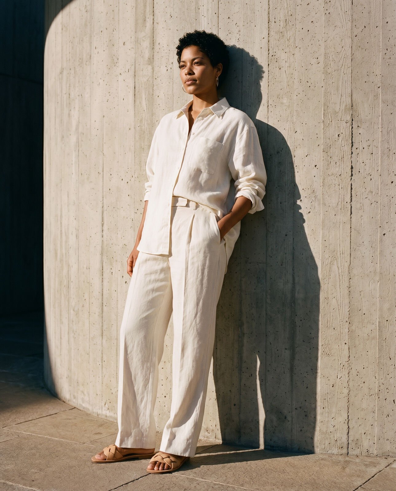 A woman in a cream linen shirt and trousers leaning against a concrete wall in late sun, from the MARESIA campaign