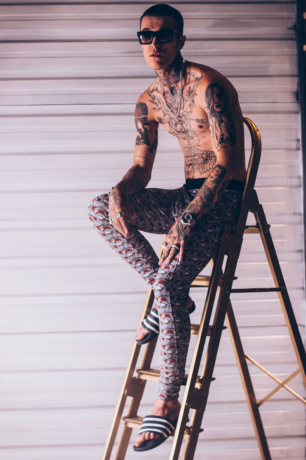 A tattooed model in printed underwear and sunglasses sitting on a gold ladder